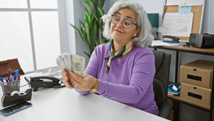 A mature woman examines polish zloty bills in an indoor office setting, depicting finances,...