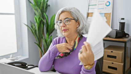 Mature woman fanning herself with paper in a modern office setting, expressing discomfort or heat.