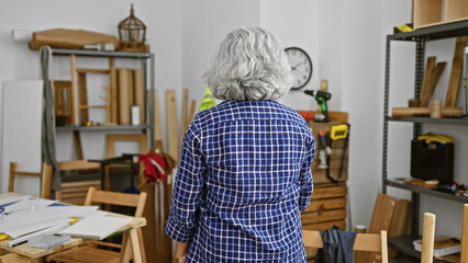 A grey-haired woman in a checkered shirt stands contemplatively in a cluttered woodworking workshop.