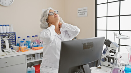 Naklejka premium Mature woman scientist with grey hair experiencing neck pain in a laboratory setting.