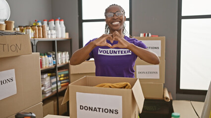 Smiling woman wearing glasses and volunteer shirt forming heart gesture in donation center with...