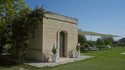 Obraz premium A rustic stone building surrounded by greenery and outdoor seating areas under canopies in the serene countryside of puglia, italy on a sunny day.
