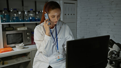 A young, caucasian woman in a laboratory, wearing a lab coat and talking on the phone while using a computer, portraying a professional healthcare setting