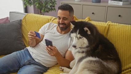 A smiling young hispanic man with a credit card and phone sitting beside a husky on a yellow couch indoors