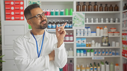 Confident hispanic man with glasses standing thoughtfully in a pharmacy full of health products. © Krakenimages.com