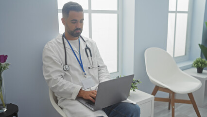 Hispanic male doctor in white lab coat using laptop in a bright clinic office, embodying professionalism and healthcare.