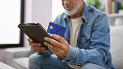 Smiling middle-aged man with gray hair, using a tablet and holding a credit card indoors.