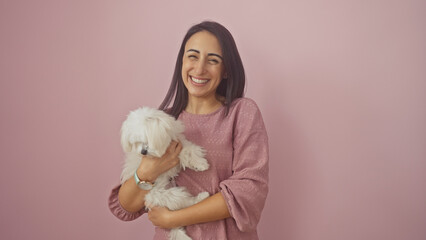 A happy hispanic adult woman in a pink blouse holding a white bichon maltese dog against a pink background