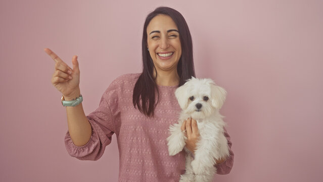 A cheerful young hispanic woman points to the side while holding a white bichon maltese dog against a pink isolated background.