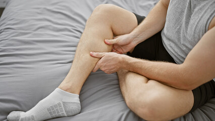 A young man sitting on his bed at home, touching his calf in discomfort, suggesting a leg pain or muscle cramp. © Krakenimages.com