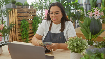 Obraz premium Middle-aged woman counts romanian currency in a flower shop filled with green plants and flowers, depicting a small business atmosphere.