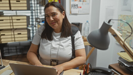 A smiling mature hispanic woman detective in an office with a badge necklace using a computer,...