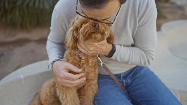 A middle-aged hispanic man gives a loving kiss to his pet poodle while sitting outdoors in an urban park setting.