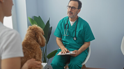 A hispanic man in veterinary scrubs consults a tablet in a clinic, facing a poodle and its owner.