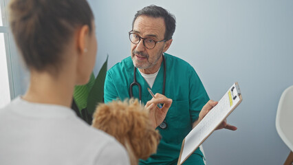 A hispanic man in scrubs consults with a customer holding a poodle in a veterinary clinic room.