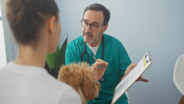 A hispanic man in scrubs consults with a customer holding a poodle in a veterinary clinic room.
