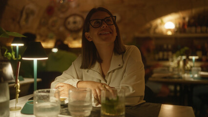 A young hispanic woman enjoying her time at an indoor restaurant in italy, surrounded by a cozy and warm ambiance with glasses and lamps on the table.