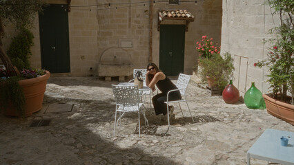 A young, hispanic, beautiful woman sits at an outdoor cafÃ© in the old town of matera, basilicata, italy, surrounded by rustic stone buildings and potted plants on a sunny day.