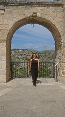 Fototapeta na wymiar A young hispanic woman in black attire stands at a viewpoint in the old town of matera, basilicata, italy, framed by a historic stone arch with a scenic background of hills and clear blue skies.