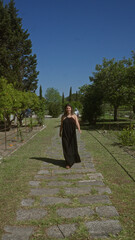 Beautiful young hispanic woman walks outdoors at a picturesque villa in puglia, italy, surrounded by lush greenery and a clear blue sky.