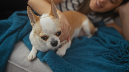 A young woman relaxes at home, gently touching her pet chihuahua on a couch with a blue blanket.