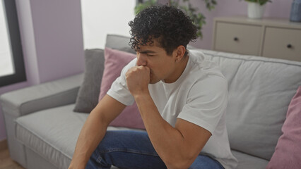A pensive young hispanic man sits thoughtfully in his well-lit living room, embodying a casual and modern domestic setting.