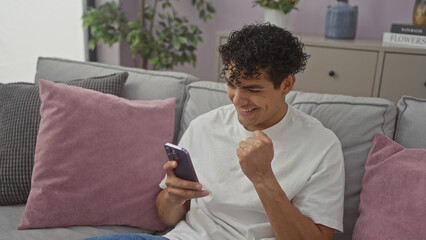 A cheerful hispanic man celebrates success while using a smartphone in a cozy living room setting.