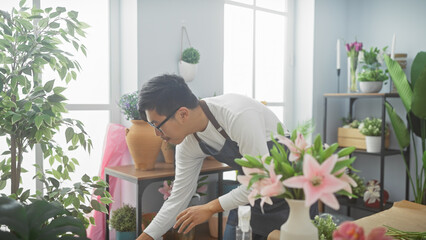 A young asian man arranges flowers in a bright, green-filled interior flower shop.