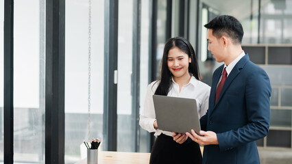 Two business professionals in formal attire discussing work on a laptop in a modern office setting with large windows.
