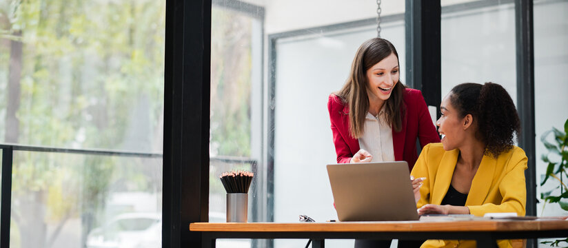 Two professional women working together on a laptop in a contemporary office setting with large windows and natural light.