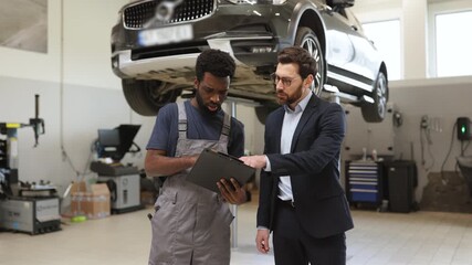 Mechanic explaining car repair details to customer in modern auto shop. Professional interaction between mechanic and client in vehicle service center with car on lift.