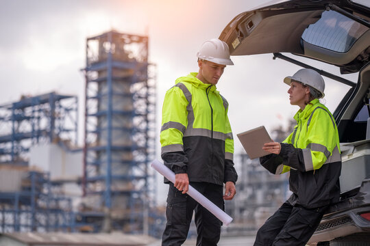 Engineers wear uniform standing near the open trunk of a vehicle hand holding blueprint paper show, survey inspection work plant site use tablet see detail of work with oil refinery background.