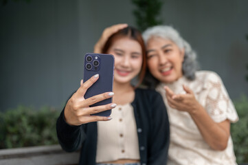 Fototapeta premium A lovely Asian granddaughter and her grandma are sitting on a bench and taking a selfie together.