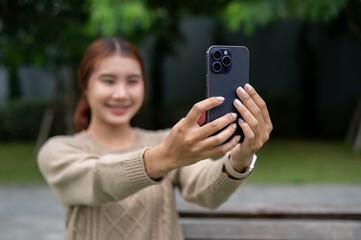 A positive Asian woman is taking a selfie or talking on a video call on a bench in a park.