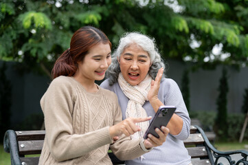 Fototapeta premium A caring, happy Asian granddaughter is showing a video on her smartphone to her grandma, in a park.