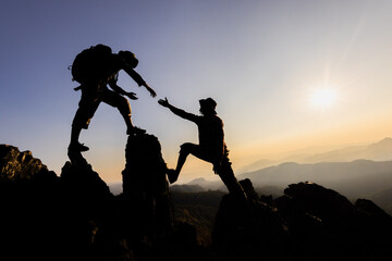 Silhouette of two mountaineers offer helping hand on a rock ridge at sunrise above a valley....