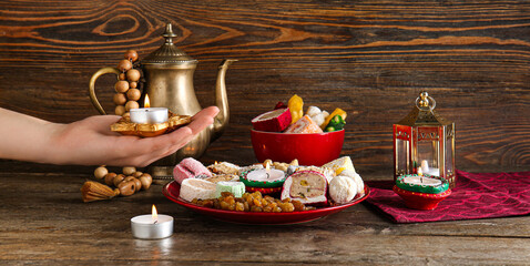 Female hand with diya lamps, candles, beads, teapot and plates of different treats on brown wooden background. Divaly celebration