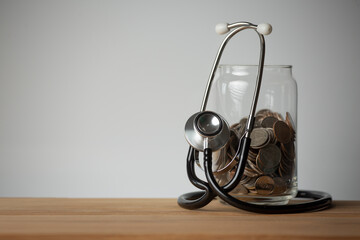 Concepts for checking health, finances and saving money. Coins in a glass jar, Stethoscope, on a wooden table.