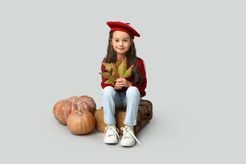Cute little girl in stylish clothes with autumn leaves, pumpkins and suitcase on grey background