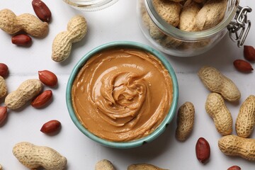 Tasty peanut butter in bowl and groundnuts on white tiled table, flat lay