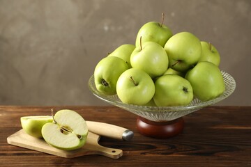 Glass vase with fresh green apples, cutting board and knife on wooden table against gray background
