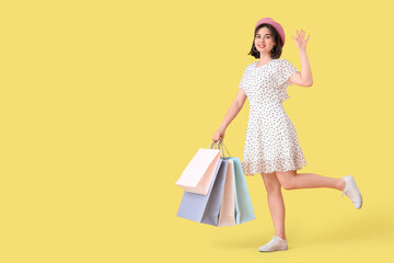 Portrait of fashionable young woman in beret with shopping bags waving hand on yellow background