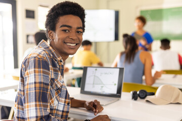 Smiling teenage boy using laptop in classroom, studying with classmates in high school