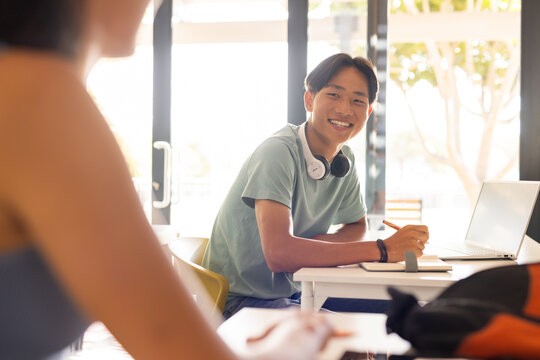 Smiling teenage boy with headphones studying at high school desk with laptop and notebook