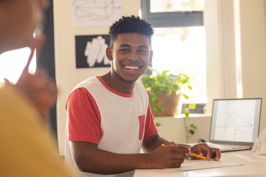 In high school, smiling teenage boy writing in notebook with laptop on desk