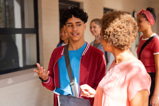 Teenager talking with teacher, holding tablet, walking in high school hallway
