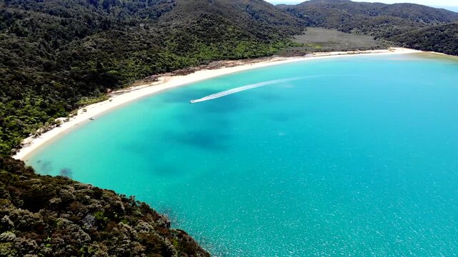 Aerial view of a speed boad plying the clear blue waters of Able Tasman and comes to a stop at a white sand beach surrounded by native bush. At Able Tasman National Park in New Zealand.