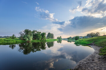 A winding river flows through a serene landscape, surrounded by vibrant green foliage. The sky is a canvas of fluffy clouds, their reflections mirrored in the still water.