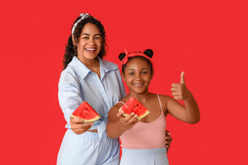 Beautiful young African-American woman and her cute daughter with pieces of fresh watermelon showing thumb-up gesture on red background