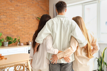 Young man with two women hugging in kitchen, back view. Polyamory concept